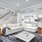 A large room with a white sectional couch and a grey couch next to a staircase with a kitchen in the background.