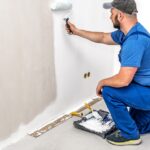 A man in a gray hat, blue T-shirt, and blue overalls kneels down to paint a wall white with a roller.