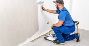 A man in a gray hat, blue T-shirt, and blue overalls kneels down to paint a wall white with a roller.