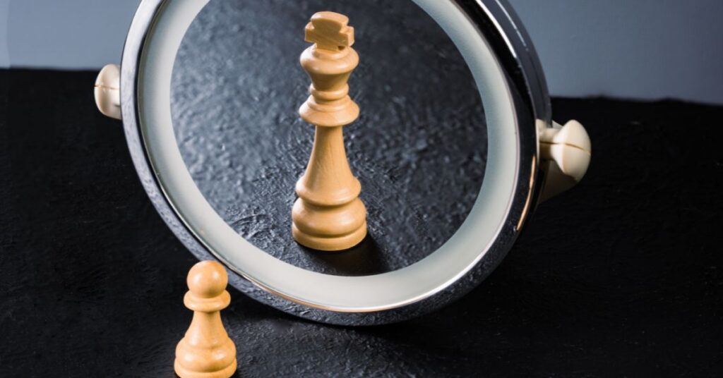 A wooden chess pawn placed in front of a small circular mirror that is reflecting back a chess king.