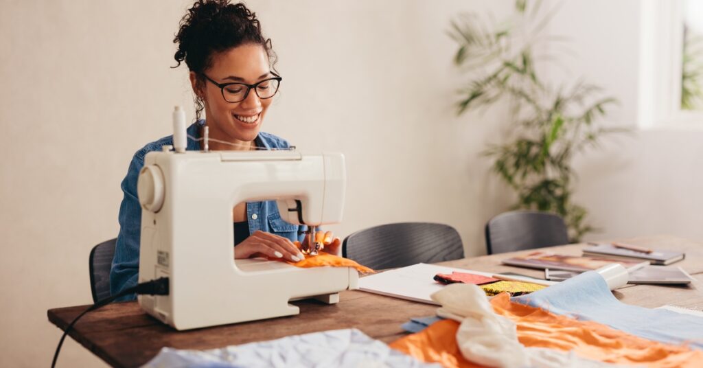 A smiling woman wearing black glasses and a denim shirt uses a white sewing machine to sew orange fabric.