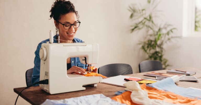 A smiling woman wearing black glasses and a denim shirt uses a white sewing machine to sew orange fabric.