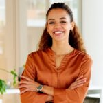 A woman wearing an orange silk blouse and a black watch smiles while standing next to a potted plant.