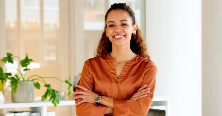 A woman wearing an orange silk blouse and a black watch smiles while standing next to a potted plant.
