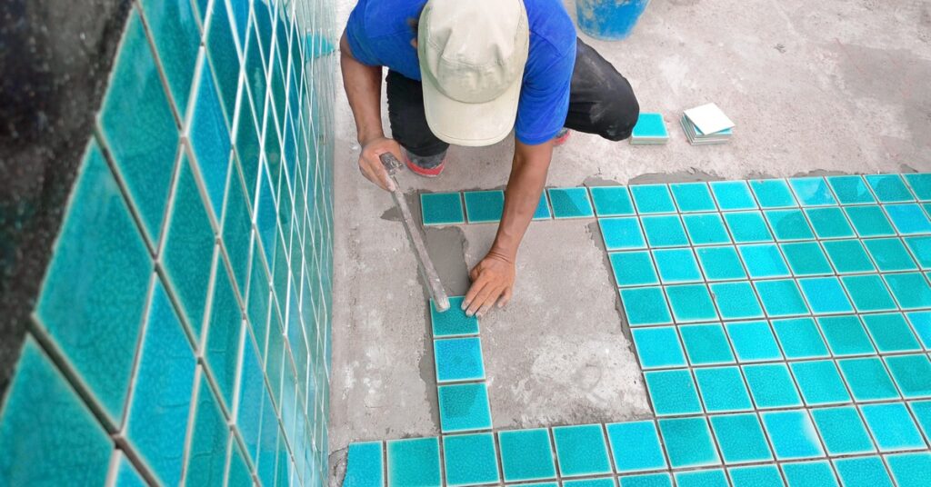 A worker tiling a pool with blue-green square tiles, using a trowel, surrounded by buckets, tools, and stacked tiles.