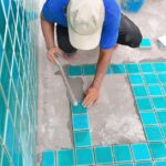 A worker tiling a pool with blue-green square tiles, using a trowel, surrounded by buckets, tools, and stacked tiles.