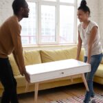 A man and a woman stand on opposite ends of a white coffee table by an L-shaped couch. They're holding the table in the air.