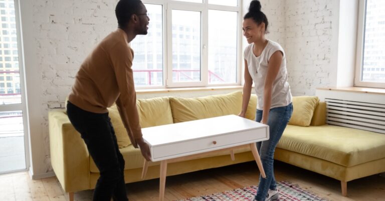 A man and a woman stand on opposite ends of a white coffee table by an L-shaped couch. They're holding the table in the air.