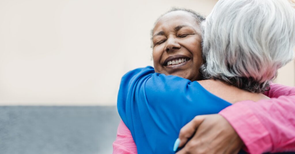 Two women hug each other while standing in a hallway of a building. One wears a pink shirt and the other wears blue.