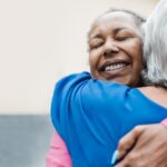 Two women hug each other while standing in a hallway of a building. One wears a pink shirt and the other wears blue.