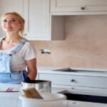An older woman wearing denim overalls looks up to her kitchen. On the table are paint swatches and paint cans.