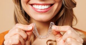 A close-up of the lower half of a woman's face. She's smiling and holding a plastic teeth aligner mold.