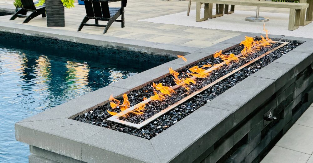 A rectangular fire feature set up next to a swimming pool and an outdoor kitchen with a table and bench seats.