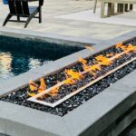 A rectangular fire feature set up next to a swimming pool and an outdoor kitchen with a table and bench seats.
