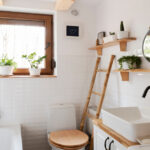A small white bathroom with tub, toilet, and sink features natural wood shelving, a ladder, and green plants.