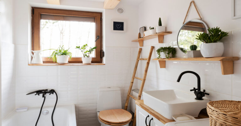 A small white bathroom with tub, toilet, and sink features natural wood shelving, a ladder, and green plants.