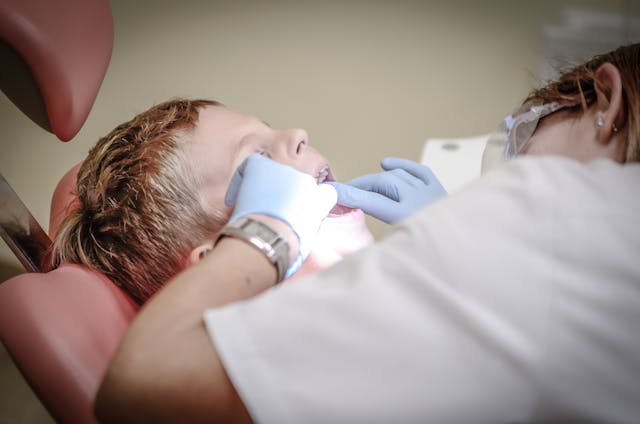 Female dentist in white scrubs and gloves examines a young boy’s teeth as he reclines in a dental chair at a clinic