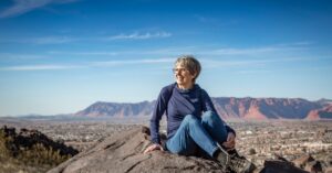 An older woman in glasses sits on a rocky overlook and smiles with a desert town, red mountains, and blue sky behind her.