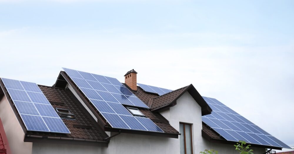 Solar panels covering multiple sections of a sloped brown shingle roof on a white house under a clear blue sky.