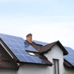Solar panels covering multiple sections of a sloped brown shingle roof on a white house under a clear blue sky.