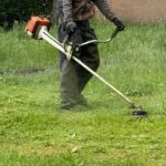 Person uses a string trimmer to cut overgrown grass in a lush green yard, clearing edges with steady precision.