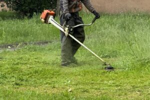 Person uses a string trimmer to cut overgrown grass in a lush green yard, clearing edges with steady precision.