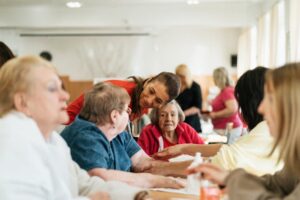 Caregiver in a red shirt speaks with a group of seniors seated around a table in a bright community room.