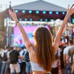 A young woman in a tank top and jeans raises her arms while standing in a crowd facing an outdoor concert stage.