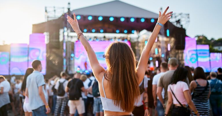 A young woman in a tank top and jeans raises her arms while standing in a crowd facing an outdoor concert stage.