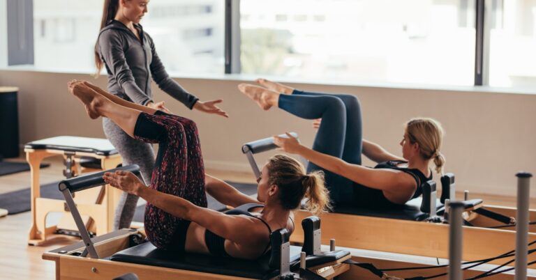 A Pilates instructor stands between women lying on reformer machines. Their knees are bent and their feet elevated.