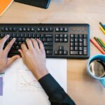 Worker typing on a black keyboard at a desk with charts and coffee, focused on getting tasks done.