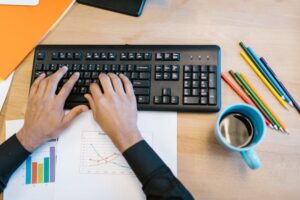 Worker typing on a black keyboard at a desk with charts and coffee, focused on getting tasks done.