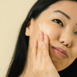 Woman with long dark hair applies a small amount of face cream to her cheek, highlighting a simple skincare step for naturally radiant skin.