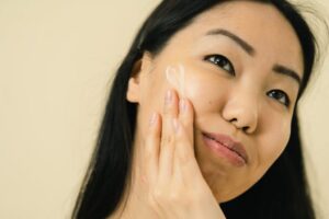 Woman with long dark hair applies a small amount of face cream to her cheek, highlighting a simple skincare step for naturally radiant skin.