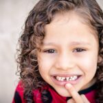 Smiling child with curly hair in a close-up portrait, showing a missing front tooth after a typical childhood dental moment.
