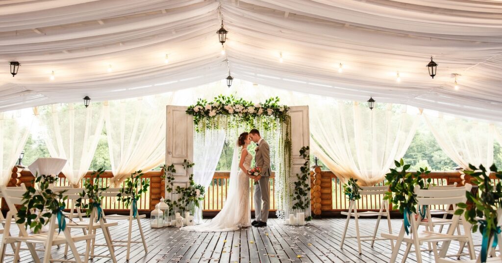 A couple standing in front of a decorative backdrop in the middle of a large wedding venue during the daytime.