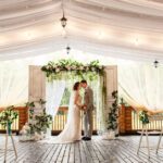 A couple standing in front of a decorative backdrop in the middle of a large wedding venue during the daytime.