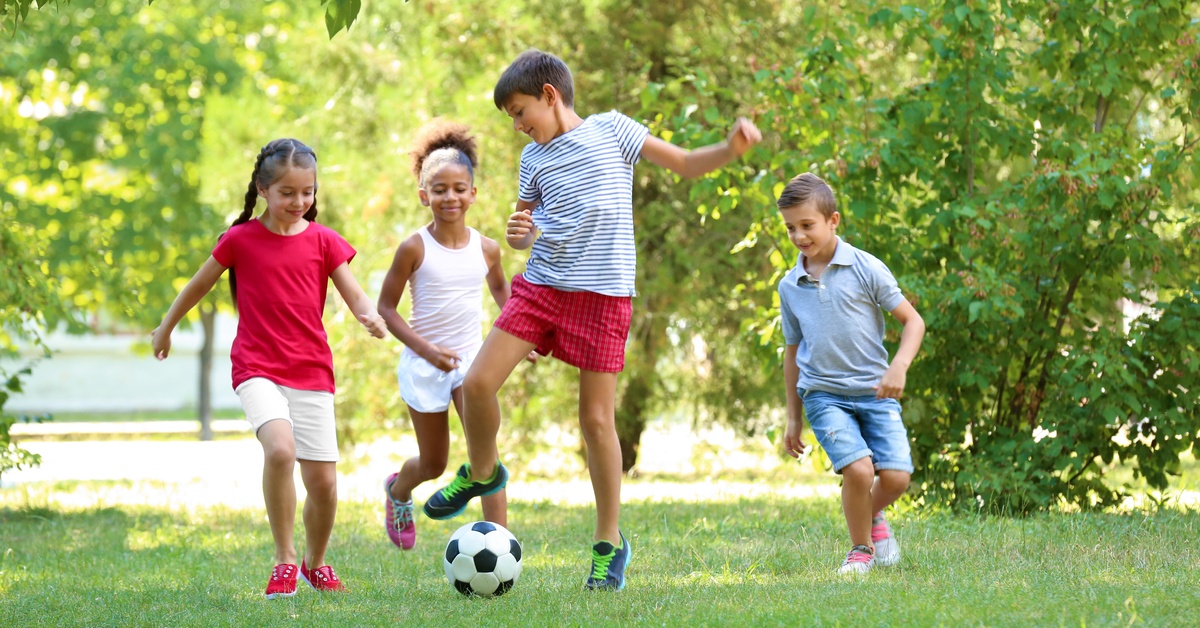 Four children running on grass near a soccer ball, with leafy trees and bushes in the sunlit background.