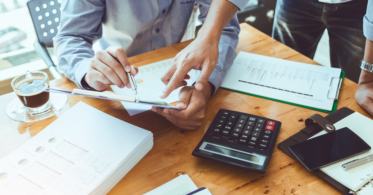 Two people review financial documents on a clipboard at a desk with a calculator, paperwork, and a cup of coffee.