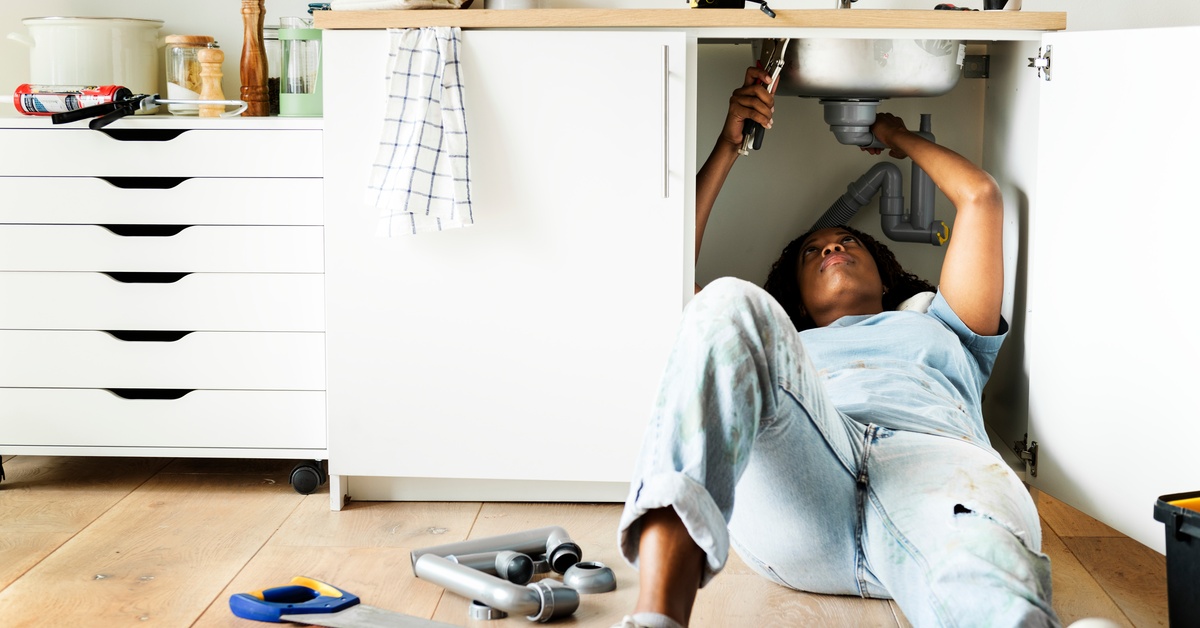 A woman lying on a wooden floor beneath a kitchen sink while reaching toward exposed plumbing under a cabinet.
