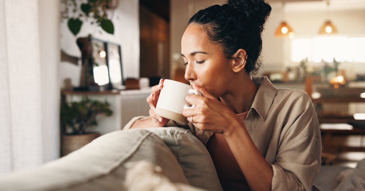 A woman sitting on a sofa and holding a white coffee mug near her mouth in a living room with pillows in front of her.