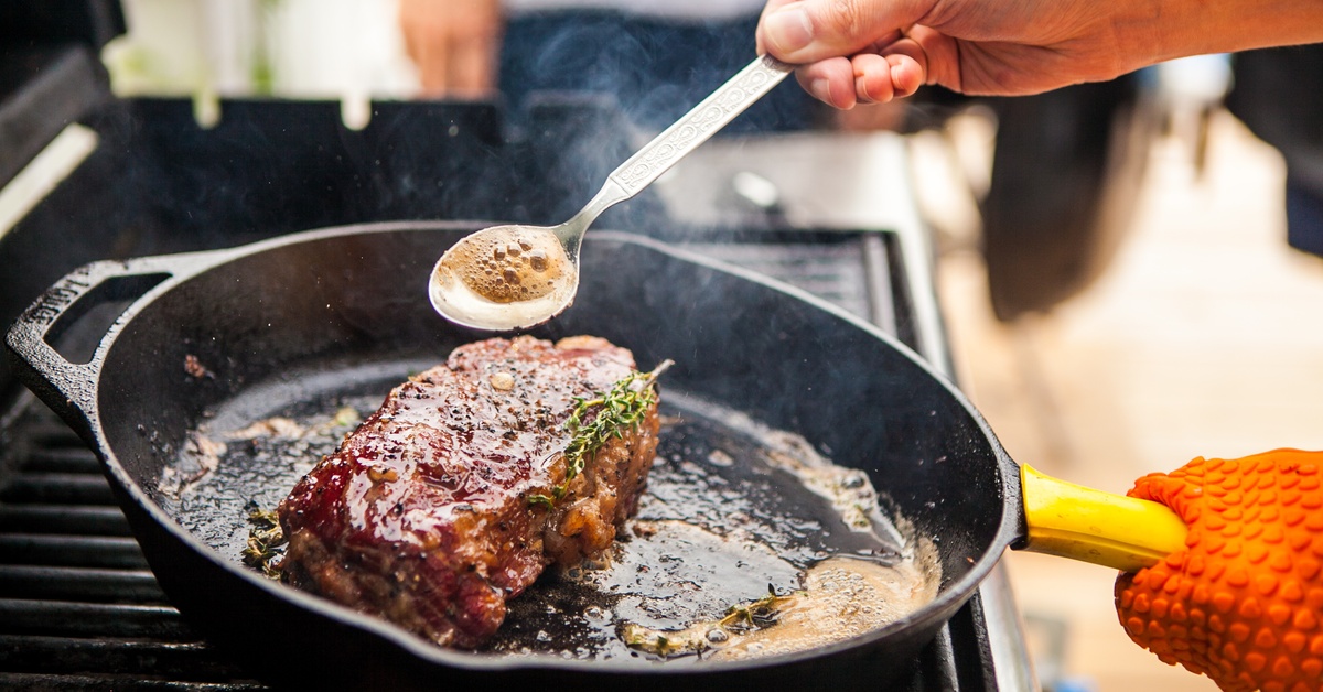 A person is cooking a steak in a cast iron skillet on a grill. They are using a spoon to baste the steak.