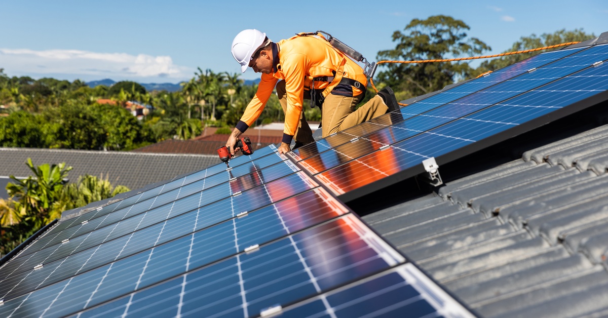 Solar installer in safety gear secures blue solar panels to a sloped residential roof under a clear sky.