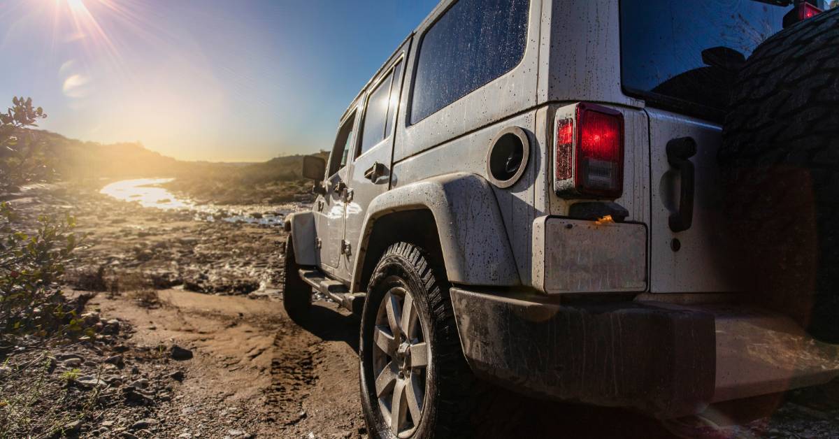 A close-up rear view from the side of a white, four-door Jeep parked on the muddy banks of a stream on a sunny day.