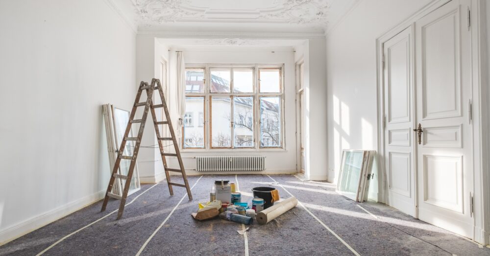 Ways To Find Properties With Renovation Potential 1 A bright white room undergoing renovation, with a wooden ladder, painting supplies, covered floors, and large windows.