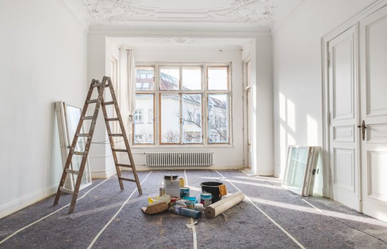 A bright white room undergoing renovation, with a wooden ladder, painting supplies, covered floors, and large windows.