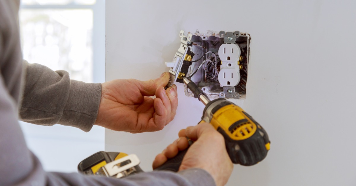 A person uses a cordless drill on the back of a light switch. The outlet is exposed, revealing wires in a wall opening.